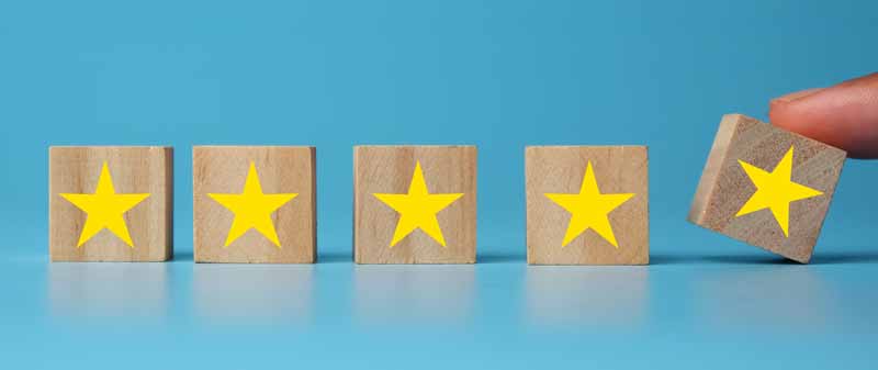 four gold stars on wooden blocks against a light blue background. a fifth gold star is being placed next to them