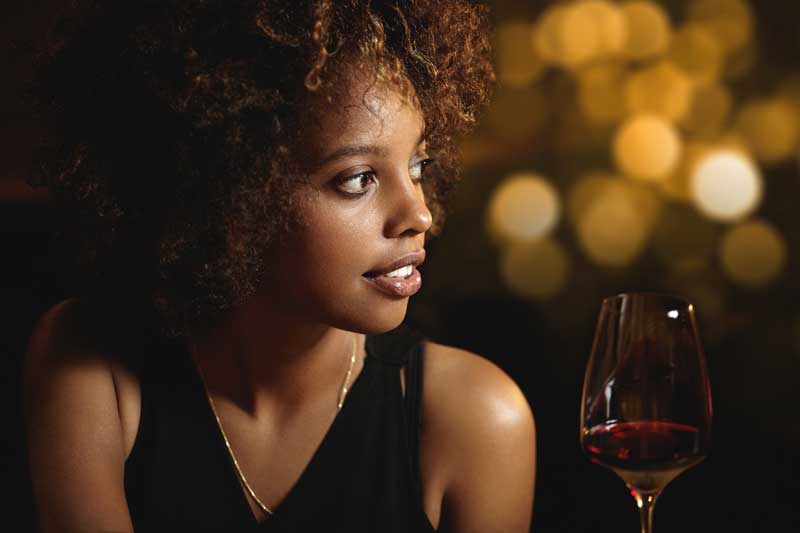profile of dark-skinned black woman with natural hair wearing a sleeveless top. Golden lights are over her shoulder and a wine glass in front of her.