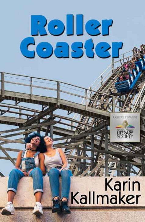 Two young women sit casually on a coral colored concrete wall in front of a wooden roller coaster featuring a car full of screaming passengers. One of the women is black with bushy natural hair, a wide smile, and is wearing overalls. The other woman is white wearing a white tank and jeans, with one arm around the other woman in an affectionate hug. Roller Coaster by Karin Kallmaker, read by Angela Dawe.