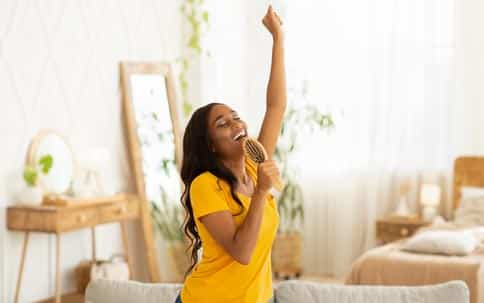 black woman with long black hair and yellow shirt thrusts one hand in the air while using hairbrush as a microphone to sing cheerfully