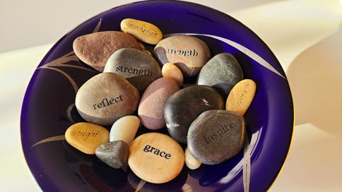 a purple bowl with gold etchings holds an assortment of reddish, orange, white, grey and black rocks. Some of the rocks are etched with empowering words like strength, insight, grace, and inspiration.