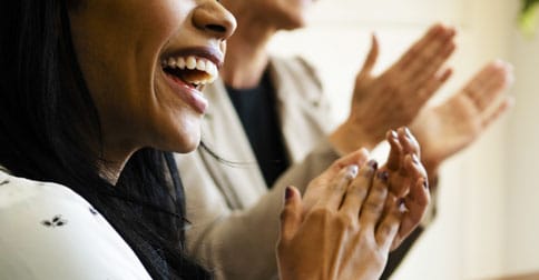 Close up of the face and hands of black business woman as she laughs and claps. Behind her another person also claps.