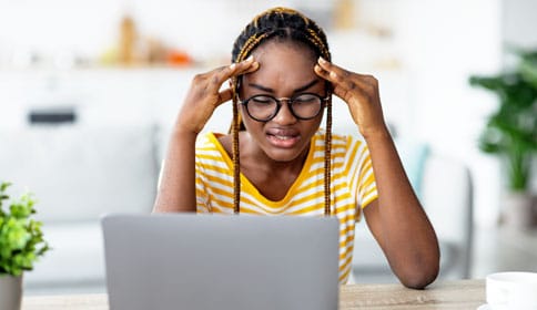 Stressed young black woman in glasses presses fingers to her temples as she looks at computer screen.