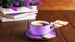 dark lavender tea mug on matching plate with cream biscuits on a dark slatted wood table. Three stacked books topped with bright lavender fill the upper left corner of the table.