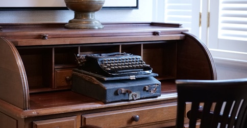 black vintage typewriter with its case sits on an antique wooden rolltop desk with cubbyholes