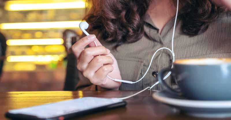 Dark-haired woman holds a headphone. In front of her in a cafe is her phone and a greyish blue cup of coffee on a matching saucer.