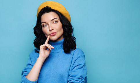 dark haired light skinned woman in blue shirt and yellow beret against plain light blue backgrounds looks likes she hatching a plan
