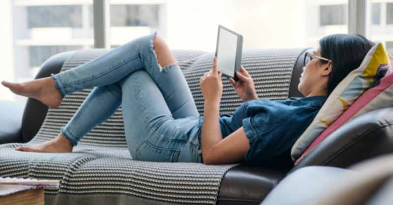 young woman reading ebook while lying comfortably on sofa at home